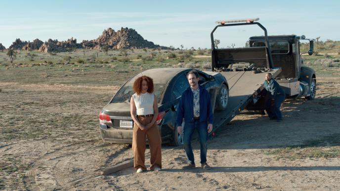 Two people sadly stand by their car that is being towed in the middle of the desert