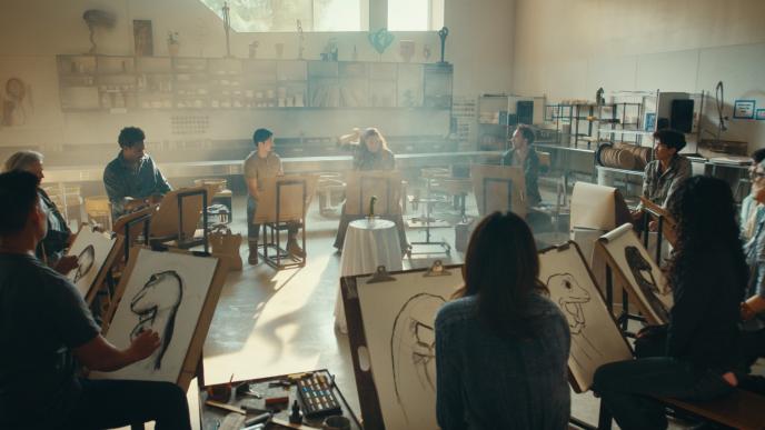 Art students circle a studio, sketching a the GEICO gecko who is posed on a stool as sunlight streams in and an instructor guides.