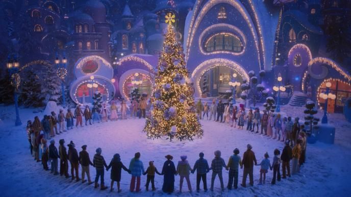 A large crowd holds hands in a snowy town square, forming a circle around a glowing Christmas tree beneath whimsical, brightly lit buildings.