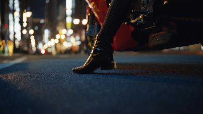 Black heeled boot stepping from a motorcycle onto a city street at night with blurred lights in background.
