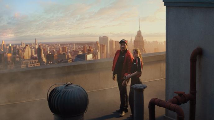 Two people in matching red jackets stand on a rooftop overlooking a city skyline at sunrise, turning toward something behind them.