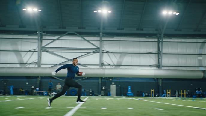 NFL star Saquon Barkley runs across an indoor field, carrying a football and wearing a blue shirt and white gloves.