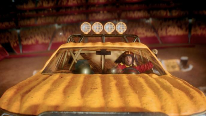 front shot of a yellow monster truck. the driver is waving their arm in victory. spectators in the background