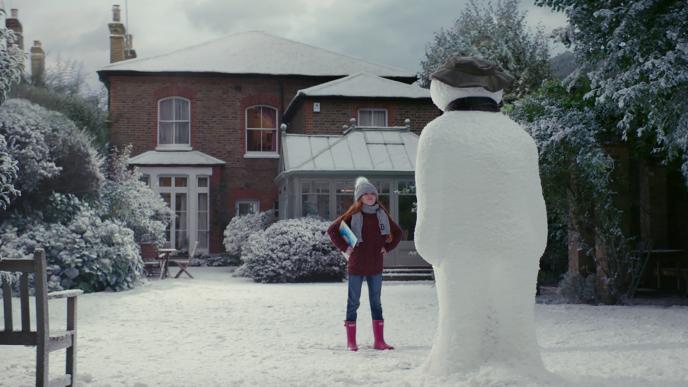 a child standing in a power position in front of a snowman, looking up at it
