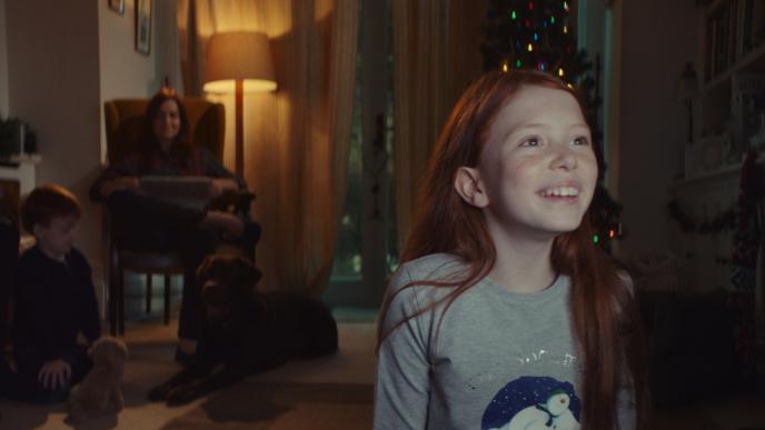 a child sitting and smiling in front of a television. the tv screen is illuminating the child's face. there is an adult and another child with a dog in the background.