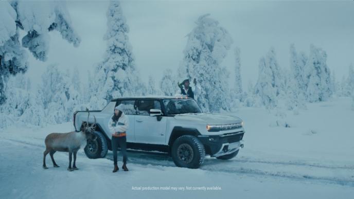 a gmc hummer ev pick up truck in the centre of a snowy terrain. there are two people standing outside each front door of the truck. there is a deer next to the person on the left. the person on the right is holding a mobile phone trying to get reception