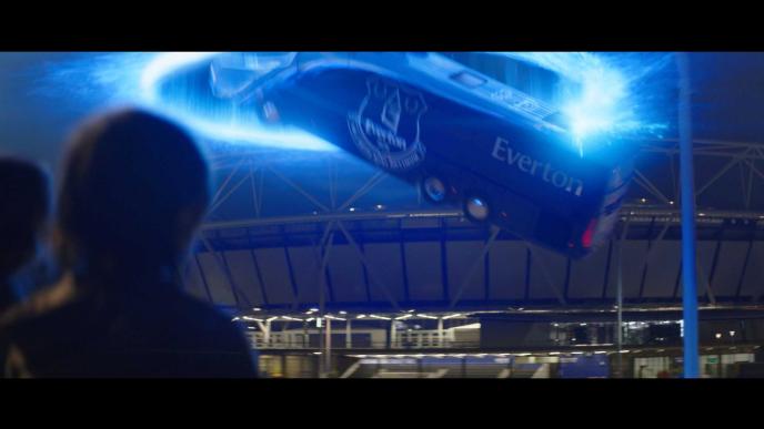two children watch on as an everton branded bus is pulled into a portal in front of a football stadium