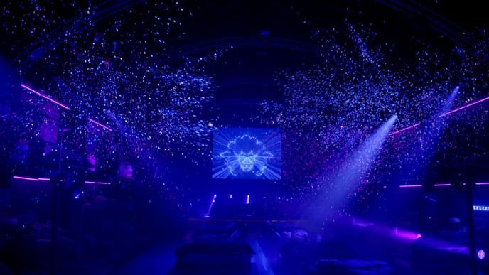 a dark dancefloor that is lit with spotlights from the right top corner. there is confetti and glitter in the air. there is a screen in the top centre that has an outline of a sheep 
