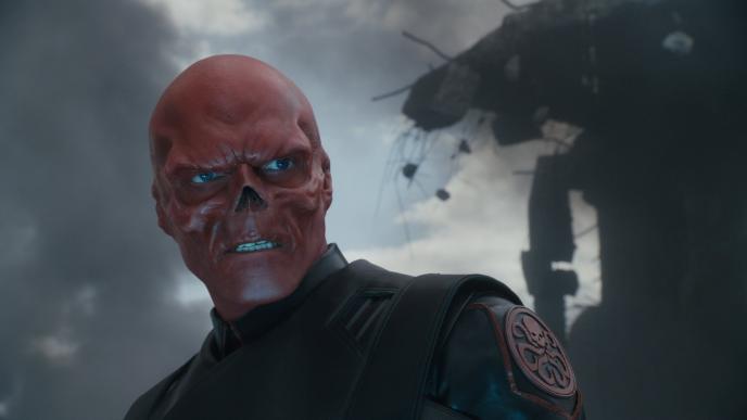 red skull character from captain marvel looking menacingly towards the right as a building in the background is falling apart