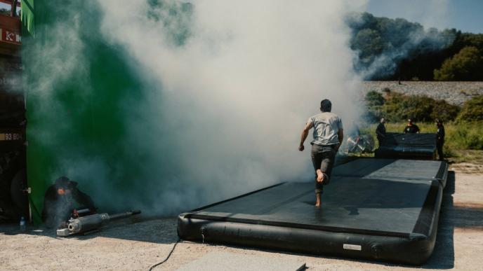 an actor running on a safety bed as a runner blows out smoke from a smoke machine in front of a green screen
