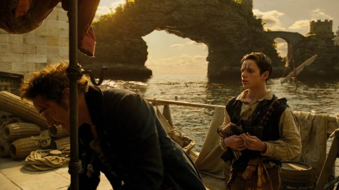 actor robert downey jr as doctor dolittle crouching and actor harry collett as tommy stubbins on a ship in front of a cavern that overlooks the sea