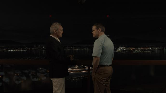 actor christopher waltz as dusan mirkovic and actor matt damon as paul safranek standing by a balcony in front of a marina full of boats and ships as a city twinkles in the background during nighttime 