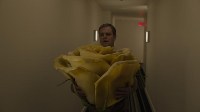 actor matt damon as paul safranek holding a giant yellow rose bud as he is walking down a building hallway