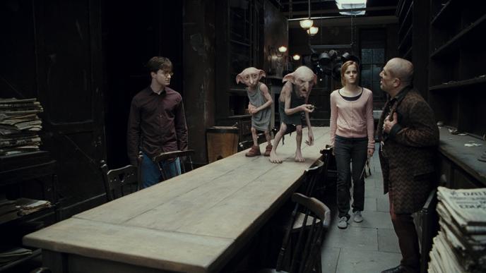 actor daniel radcliffe as harry potter, actress emma watson as hermoine granger and actor andy linden as mundungus fletcher standing around a table in a kitchen as house elves kreature and dobby stand on the table