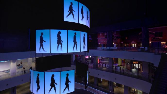 a large tower screen with silhouttes of people walking in the panels amongst a shopping mall