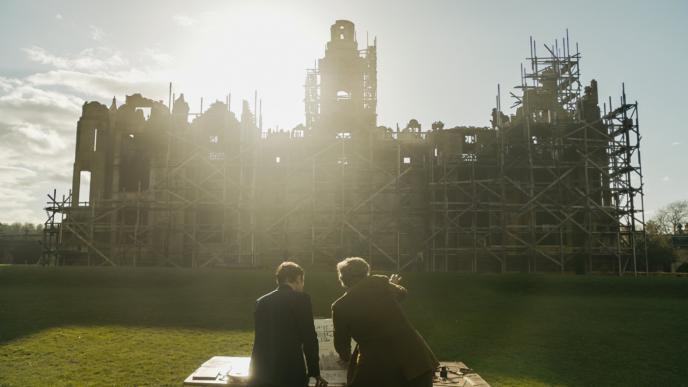 back view of two people sitting in front of a mansion covered in scaffolding as the sun beams through the building