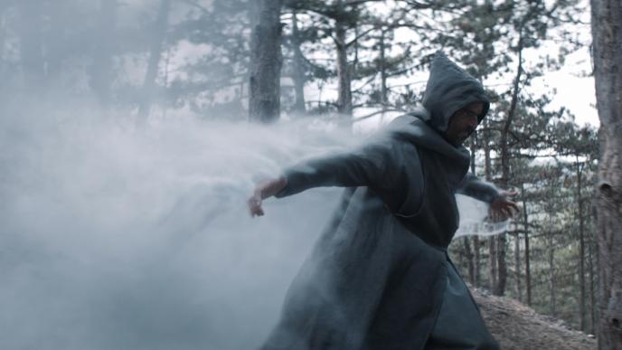 witcher character wearing a hooded cape walking through a forest as smoke comes out of his open arms