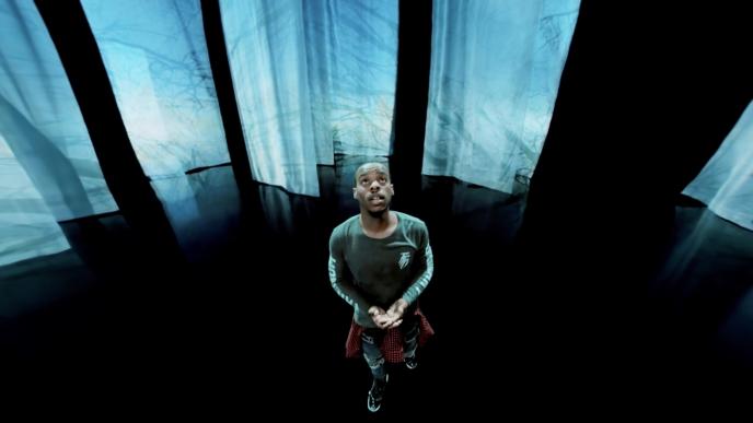 a dancer looking up with his palms open standing in front of an exhibition that has full length sheer sheets hanging from the ceiling