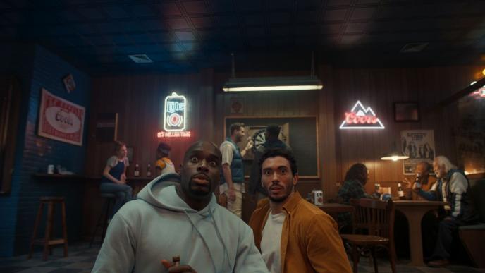 Two men sit at a bar, looking upwards at a screen