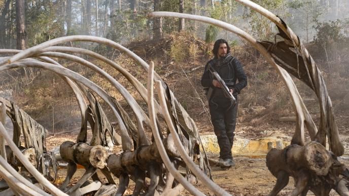 Adam Driver holds a large gun, standing near a large skeleton