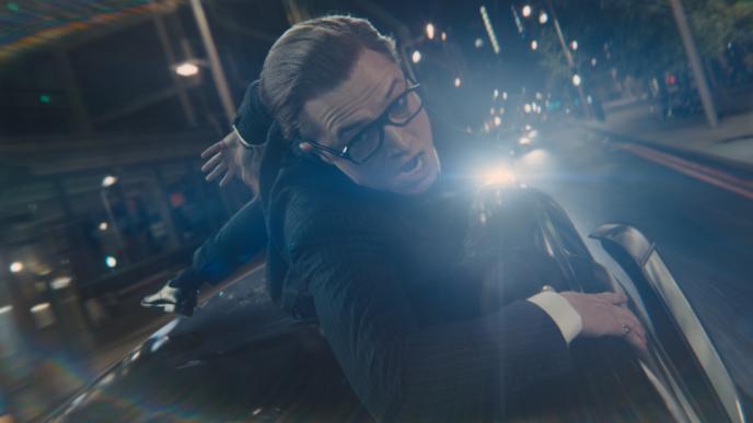 Eggsy (Taron Egerton) hangs on to the roof of a black taxi, speeding through London at night time
