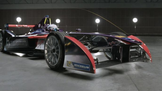 A formula E car with the front pointing towards the camera at a 45 degree angle, the car is in a large grey warehouse.