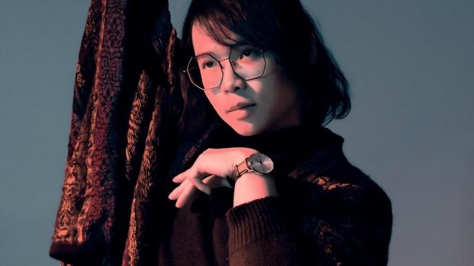 A photograph of a man holding one arm above his head and the other below his chin, he is stood against a blue background.