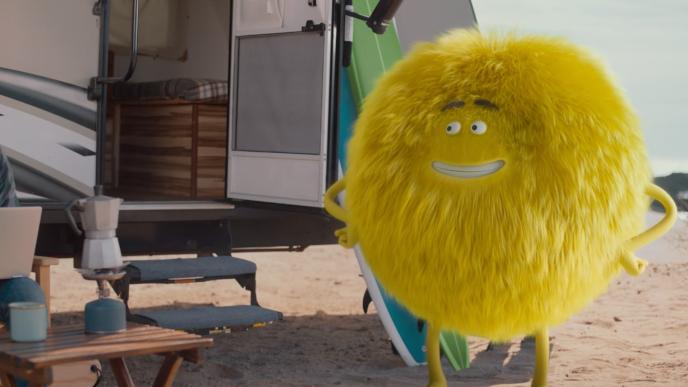 A fuzzy yellow creature stands outside an RV on the beach with a surfboard