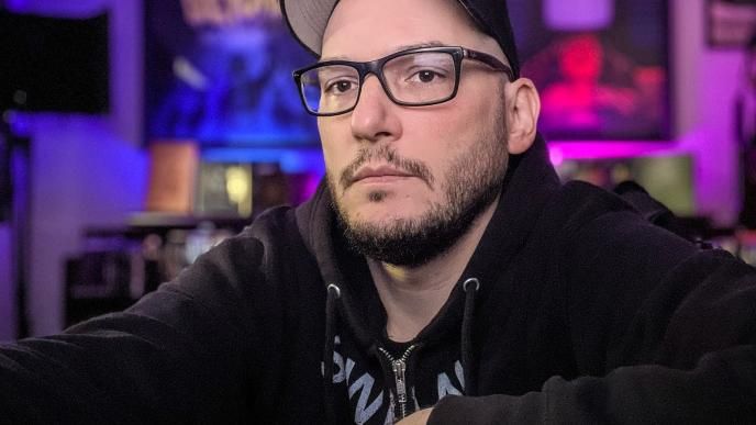 A man wearing glasses and a cap is sat in a room lit with red and blue lights, there are film and music posters in the background.