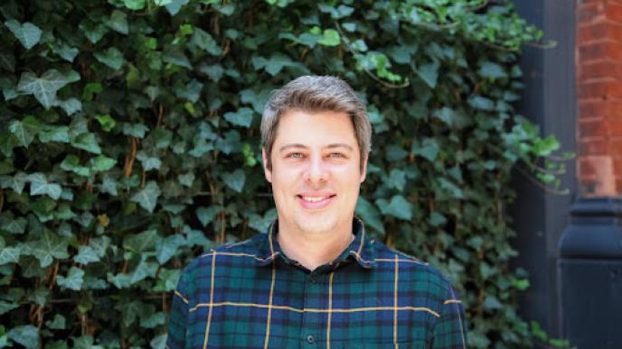 A man wearing a check shirt stood in front of a hedgerow.