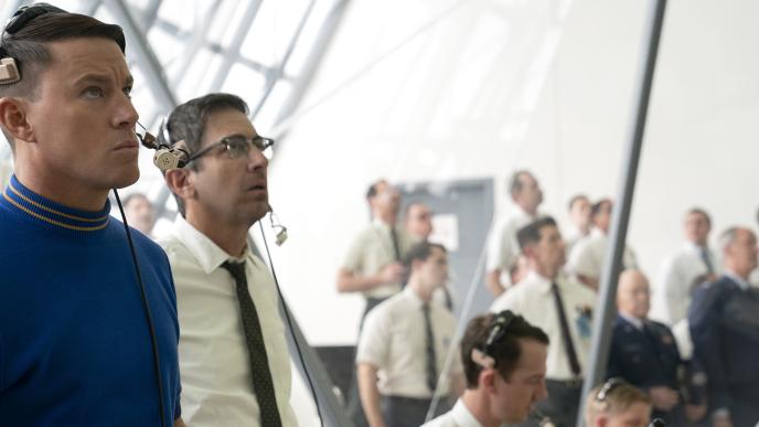 A group of men wearing shirts and ties are stood in a brightly lit room, some are wearing headsets and working at computers. 