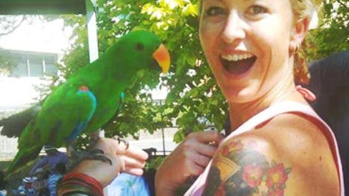 A woman with a tattoo on her shoulder holding a green parrot. 