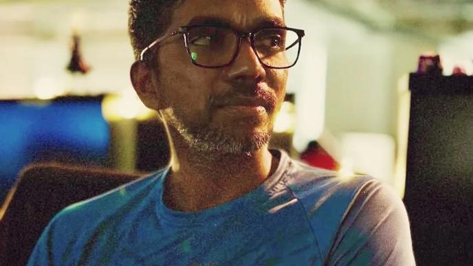 A man sat in an office wearing a blue t-shirt and glasses looking at a screen.