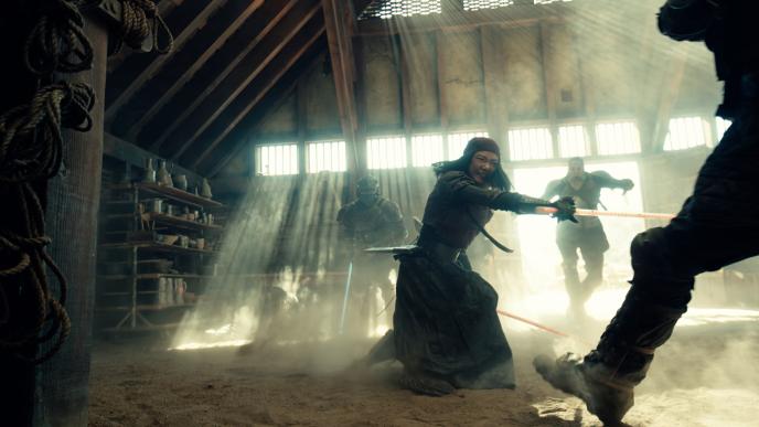A woman fighting two men with plastic stand ins for swords, they are in a dust-filled dilapidated buidling.