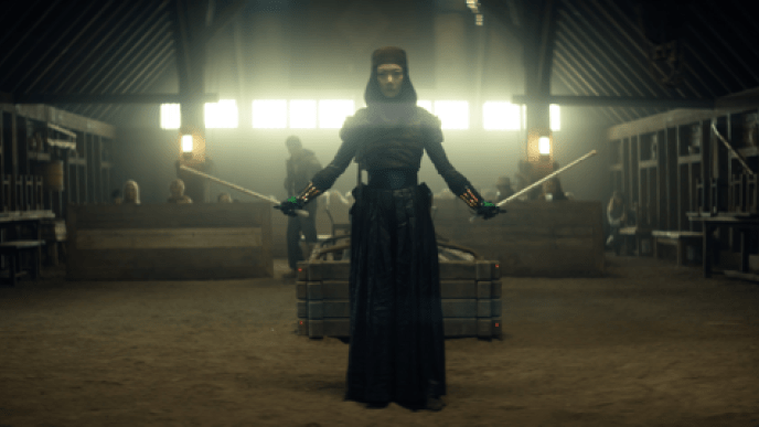 A woman holding two plastic sticks as swords, she has greenscreen gloves covering her hands. There is a light shining from behind her.