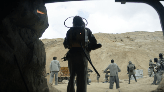 A group of people stood looking at the top of a sand dune in the background, the people are holding guns.