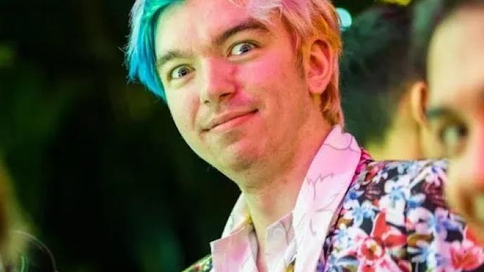 A man with blue and pink hair wearing a flowery jacket is looking at the camera.