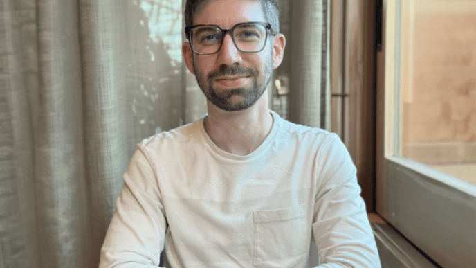 A photograph of a man wearing a cream jumper sat at table with this arms folded. 
