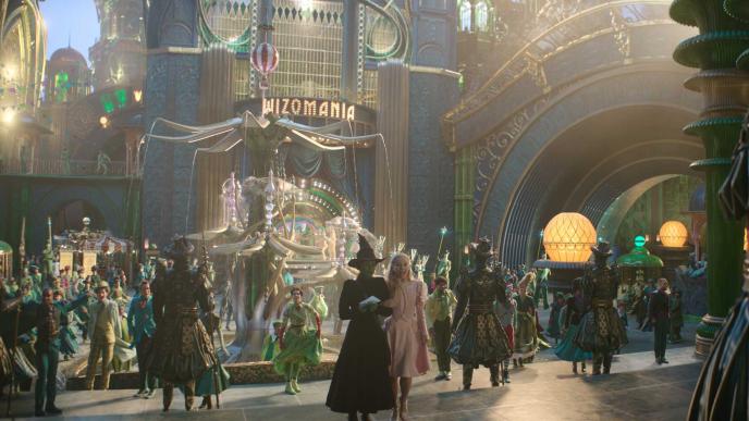 A still from the set of Wicked part one, during One Short Day. Elphaba and Glinda stand on steps, in front of a square with an elaborate fountain. 