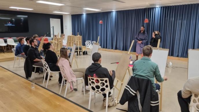  A group of people are seated with easels, observing a costumed model at the front of a well-lit room with a wooden floor and blue curtains.