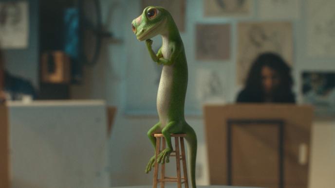 The CG animated GEICO gecko poses thoughtfully on a tiny stool in an art studio, surrounded by sketching artists and hanging drawings