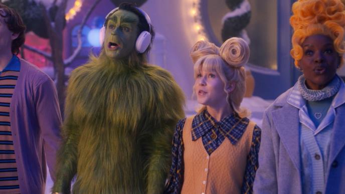 Four whimsical, brightly dressed characters—one being the Grinch with headphones—stand close together in a snowy Whoville, singing cheerfully.