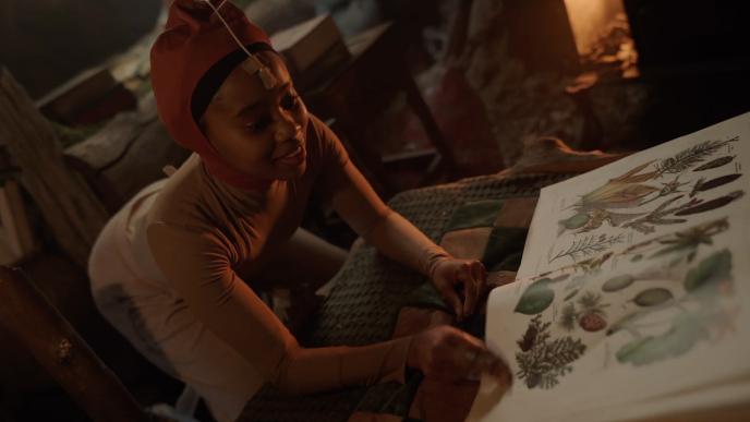 Actress N’Kone Mametja acts a physical reference for Tony Tony Chopper on set with an illustrated book of plants and insects beside warm firelight in a rustic setting.