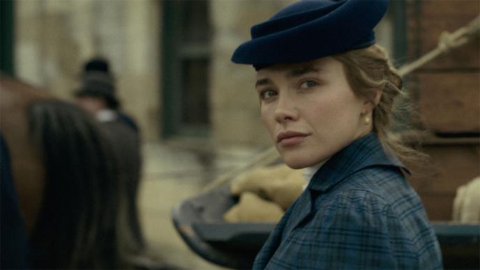 Florence Pugh as Cathy Ames in East of Eden, wearing period clothing and blue hat glances over her shoulder on a historic street, with background figures softly blurred.