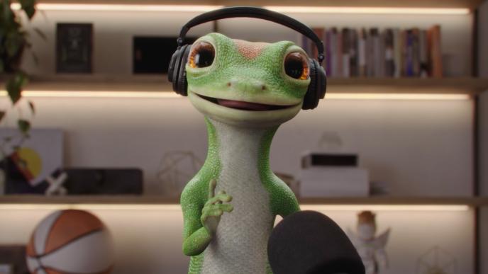 The CG animated GEICO Gecko wearing headphones speaks into a podcast microphone in a warmly lit studio with shelves, books, plants, and decorative items.
