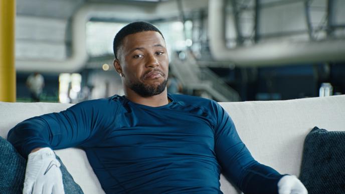 NFL star Saquon Barkley in a blue shirt sits on a couch, wearing white gloves, with an industrial interior behind him.