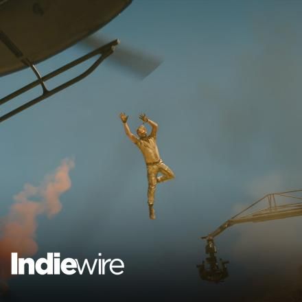 A man jumping from a crane to a helicopter through the air, there is the IndieWire logo in the bottom left corner.