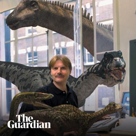 A man sat at a desk surrounded by CGI dinosaurs, some on the desk and one large on poking its head through a window.