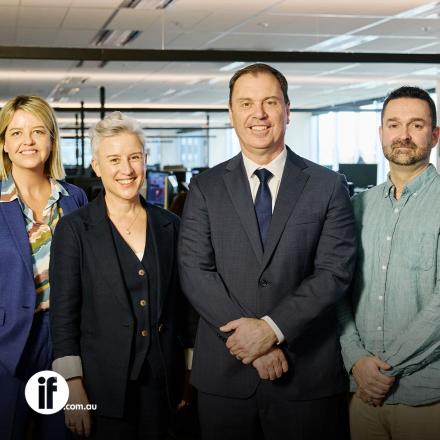 Four people stood in an office in a line wearing suits. 