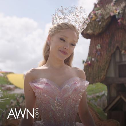 Glinda in Wicked, standing in a digitally extended Munchkinland set, with a white AWN.com logo in the lower left corner